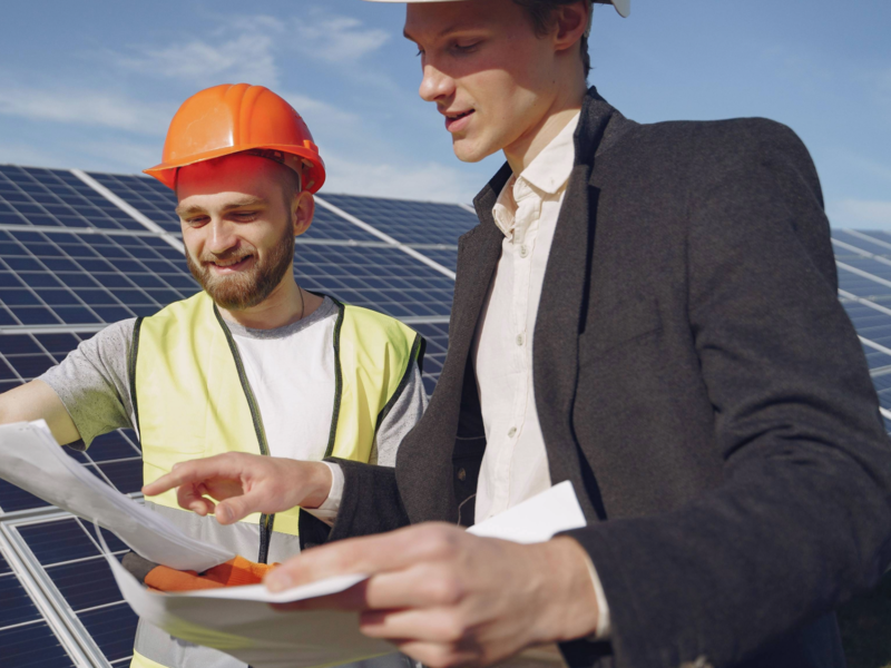 Zwei Männer in Sicherheitskleidung stehen vor Solarzellen und betrachten gemeinsam Baupläne.