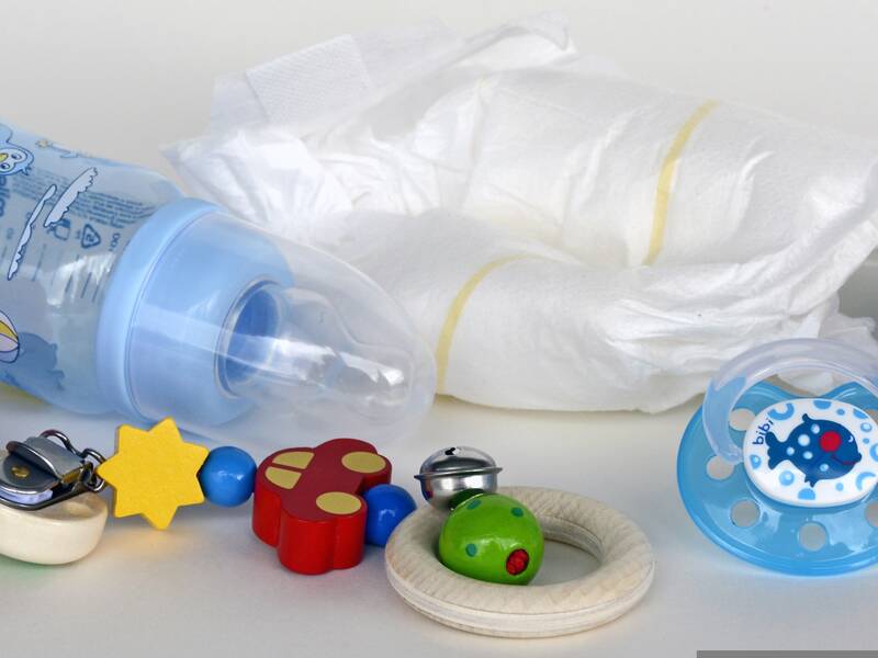 Auf dem Bild sind bunte Babyspielzeuge, eine mit blauen Motiven verzierte Babyflasche, ein Schnuller und drei Enten aus Gummi vor einem weißen Hintergrund zu sehen, wobei im Hintergrund zwei weiße Windeln liegen. Bild von Annette auf Pixabay.