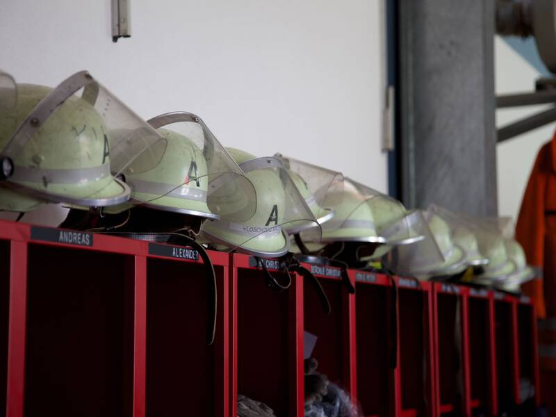 Eine Reihe von Feuerwehrhelmen mit Schutzvisieren steht ordnungsgemäß auf roten Spinden, die jeweils mit einem Namen beschriftet sind; daneben hängt eine orangefarbene Schutzkleidung.