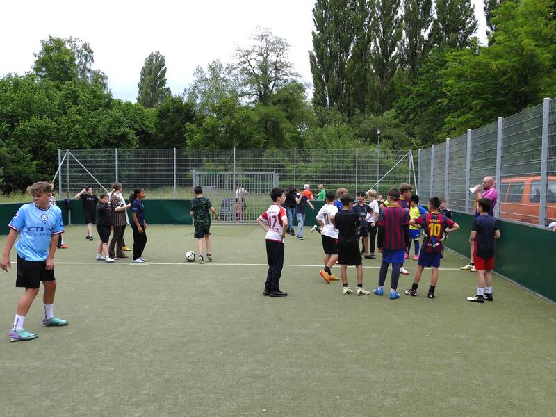 Eine Gruppe von Jugendlichen spielt auf einem umzäunten Fußballplatz, umgeben von Bäumen.