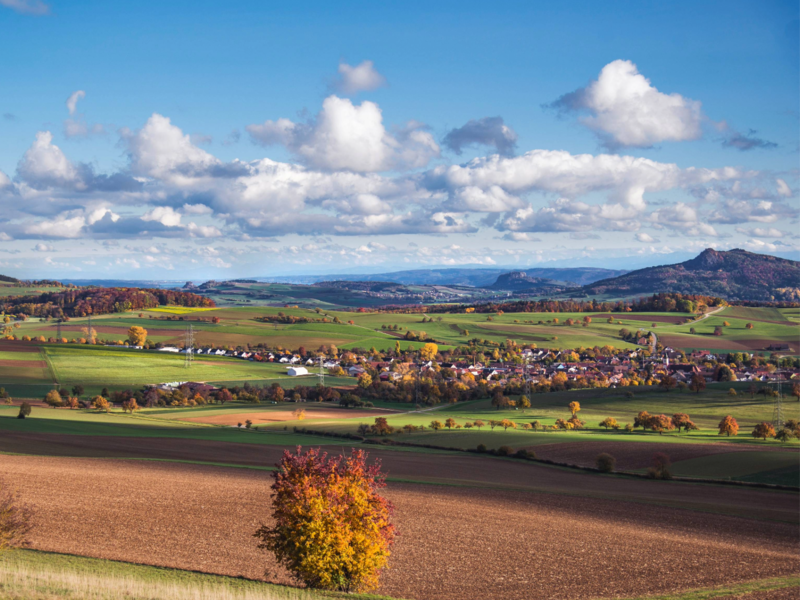 Die Landschaft zeigt weite, farbenfrohe Felder und Wiesen im Herbst, mit einem Dorf in der Mitte und den Hegau Kegeln im Hintergrund unter einem blauen Himmel mit weißen Wolken.