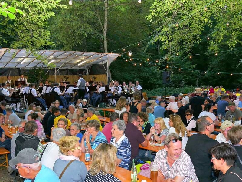 Ein großer Biergarten im Freien mit vielen Menschen, die an langen Tischen unter festlichen Lichterketten sitzen, während eine Blaskapelle im Hintergrund auf einer Bühne spielt.