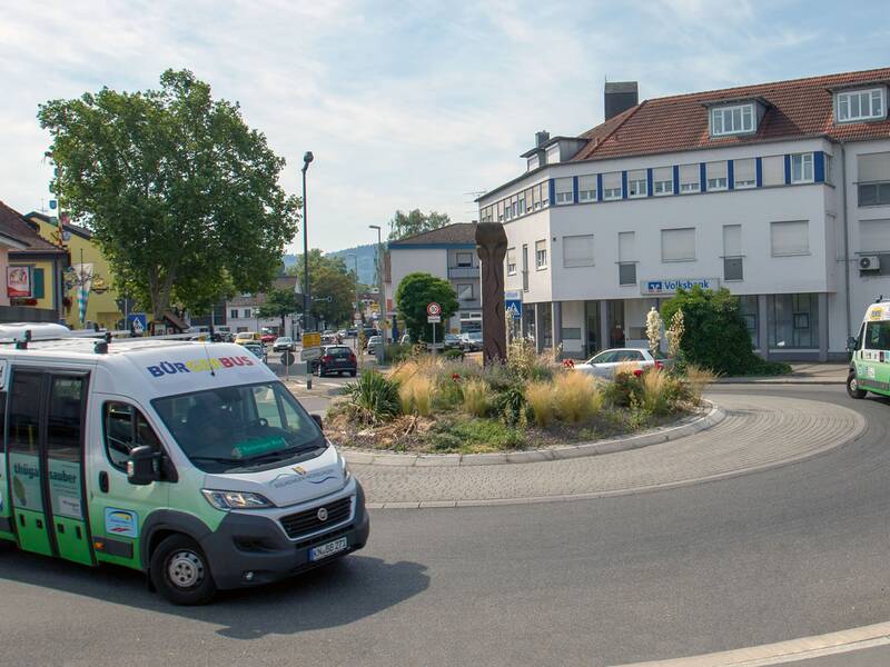 Zwei Bürgerbusse fahren durch einen Kreisverkehr, umgeben von Gebäuden und einem bepflanzten Inselbereich.