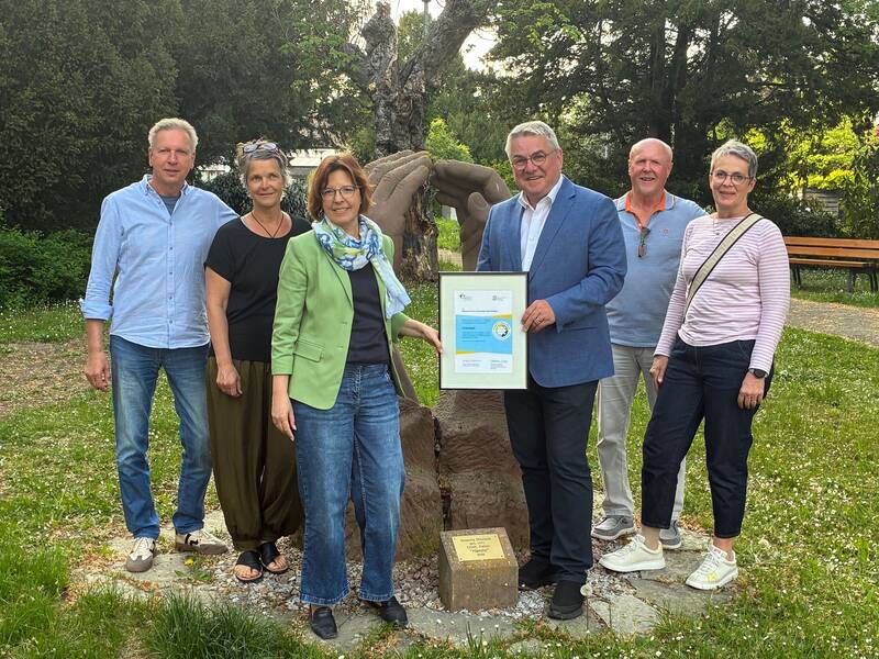 Eine Gruppe von sechs Personen steht im Freien vor einer Skulptur, und eine Person hält eine gerahmte Urkunde.