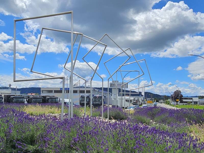 Im Vordergrund befinden sich Lavendelfelder, aus denen geometrische Rahmen wachsen, und im Hintergrund ist ein Industriegebiet unter einem bewölkten Himmel zu sehen.