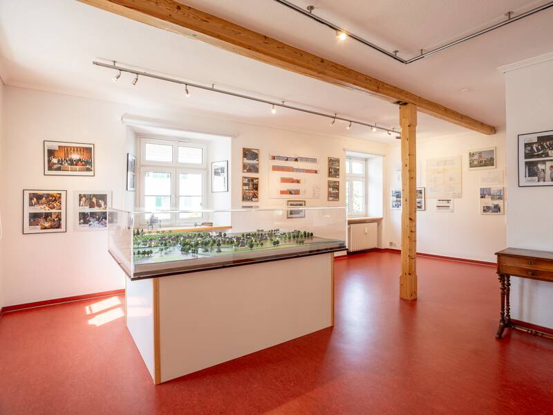 Ein Ausstellungsraum mit roten Böden zeigt ein großes Modell in der Mitte und ist von vielen Wandbildern umgeben.
