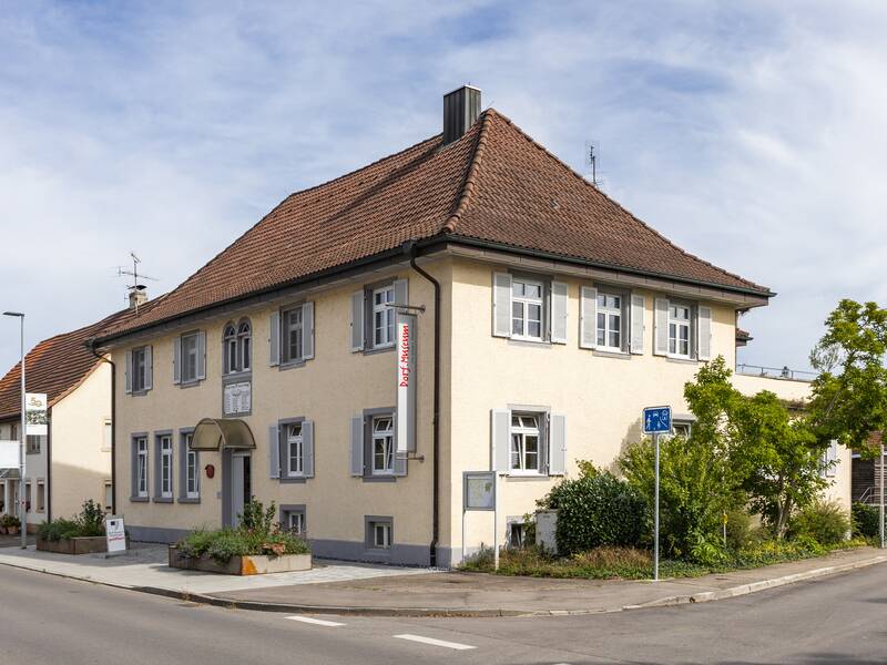 Das Bild zeigt ein zweistöckiges, helles Gebäude mit einem roten Ziegeldach, umgeben von Pflanzen, an einer Straßenecke, auf dessen Fassade ein Schild mit der Aufschrift "Dorfmuseum" hängt.