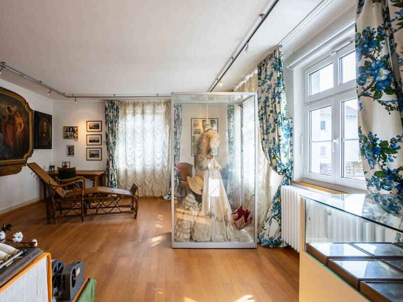 Ein heller Raum mit Holzfußboden, in dem sich eine Glasvitrine mit einem ausgestellten Kleid, ein Gemälde und ein Ruhesessel befinden, ist von reich gemusterten Vorhängen umgeben.