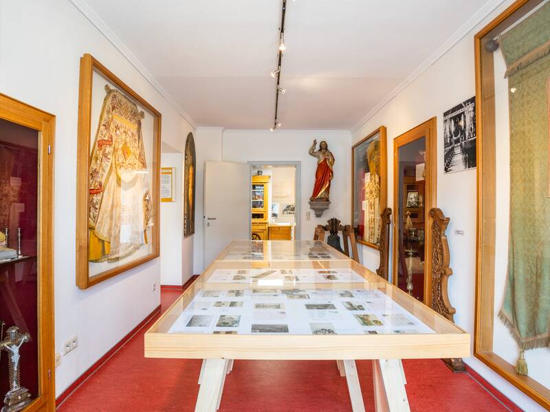 Ein schmaler Museumsraum mit roten Böden ist auf beiden Seiten mit religiösen Kunstwerken und historischen Artefakten dekoriert, und in der Mitte steht ein langer Tisch mit Dokumenten unter Glas.