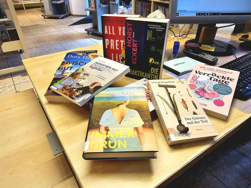Auf einem Tisch in einer Bibliothek sind sieben verschiedene Bücher ausgestellt, die neben einem Computerbildschirm liegen.