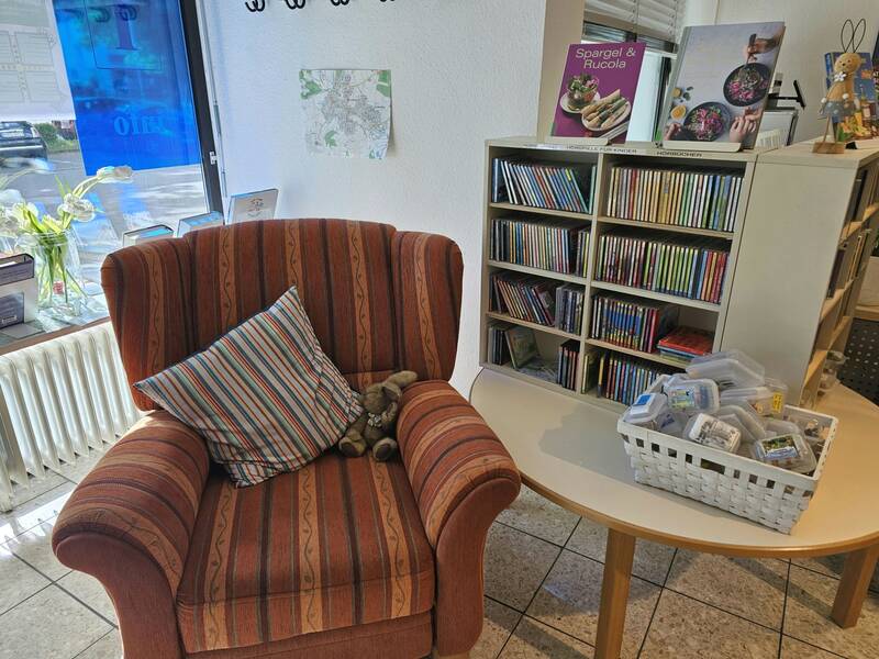 Ein gemütlicher Raum mit einem gestreiften Sessel, einem Kissen und einem Plüschtier, neben einem Tisch mit einem weißen Korb voller kleiner Boxen und einem Regal voller CDs.