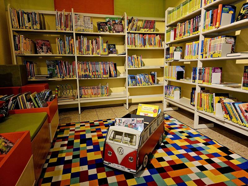 Ein gemütlicher Kinderbereich in einer Bibliothek mit bunten Regalen voller Bücher, einer Sitzgelegenheit und einem Spielzeugauto in Form eines VW-Busses auf einem farbenfrohen Teppich.