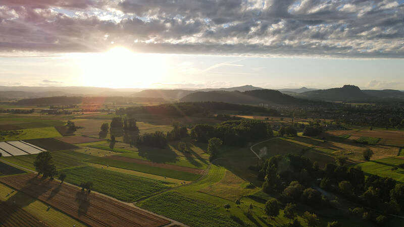 Die weitläufige Landschaft mit Feldern und Bäumen wird von der tiefstehenden Sonne beleuchtet, während Wolken den Himmel bedecken.
