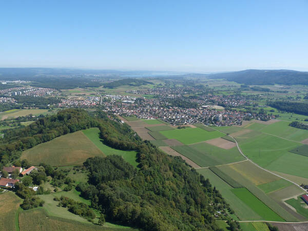 Das Bild zeigt eine weitläufige ländliche Landschaft mit grünen Feldern, Wäldern und im Hintergrund Rielasingen-Worblingen unter einem klaren blauen Himmel.