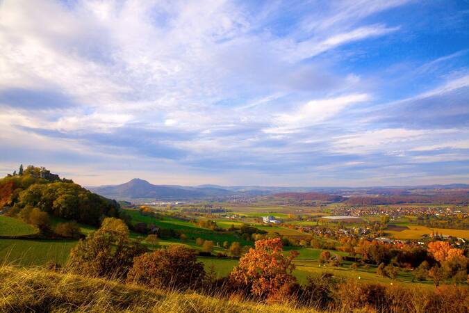 Ein weitläufiger Blick über eine herbstliche Landschaft des Hegau mit sanften Hügeln, einem kleinen Dorf und einem Berg im Hintergrund unter einem wechselhaften Himmel.