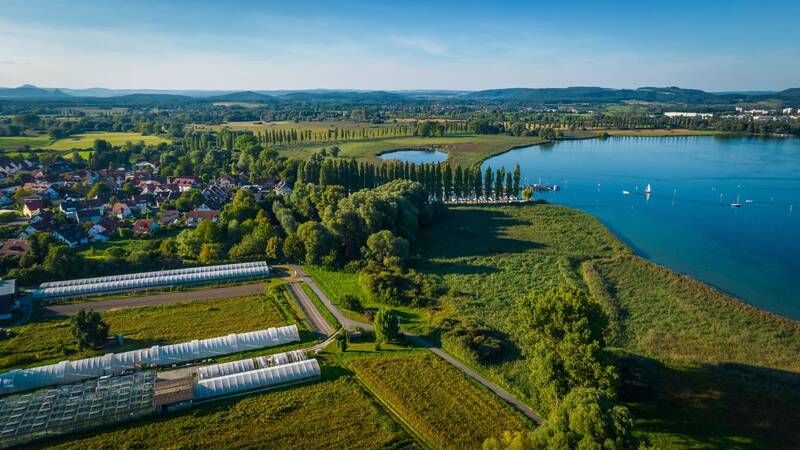 Die Luftaufnahme zeigt eine ländliche Landschaft mit Feldern, einem See mit Segelbooten und einer kleinen Siedlung rechts im Bild.