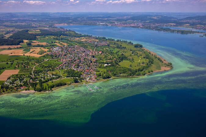 Luftaufnahme einer grünen Halbinsel mit einer Siedlung, die von einem großen See und landwirtschaftlichen Flächen umgeben ist.
