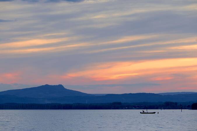 Eine Person fährt in einem kleinen Boot über den ruhigen Bodensee, während im Hintergrund die Dämmerung den Himmel in sanften Orange- und Blautönen färbt.