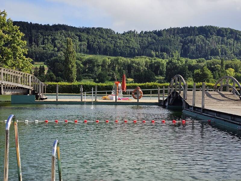 Ein Schwimmbecken mit einem Holzsteg im Vordergrund, einer Bojenlinie im Wasser und einer grünen bewaldeten Landschaft im Hintergrund.im Hintergrund.