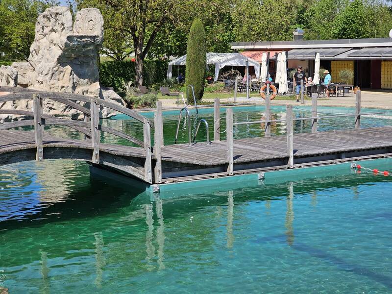 Ein Schwimmbecken mit natürlichem Wasser wird von einer hölzernen Brücke überquert, während im Hintergrund Menschen an Tischen unter Sonnenschirmen sitzen.