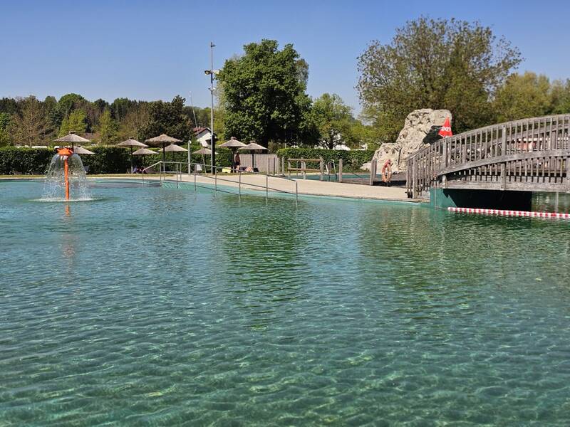 Ein Freibad mit natürlichem Wasser, einer kleinen Brücke und Sonnenschirmen im Hintergrund.