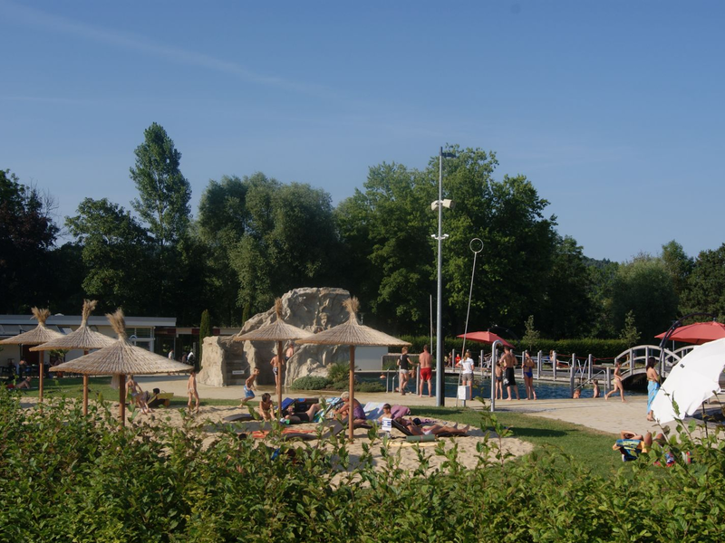 Eine sommerliche Szene zeigt einen belebten Außenbereich des Naturbades mit Sonnenschirmen, Badegästen und einer grünen Umgebung.