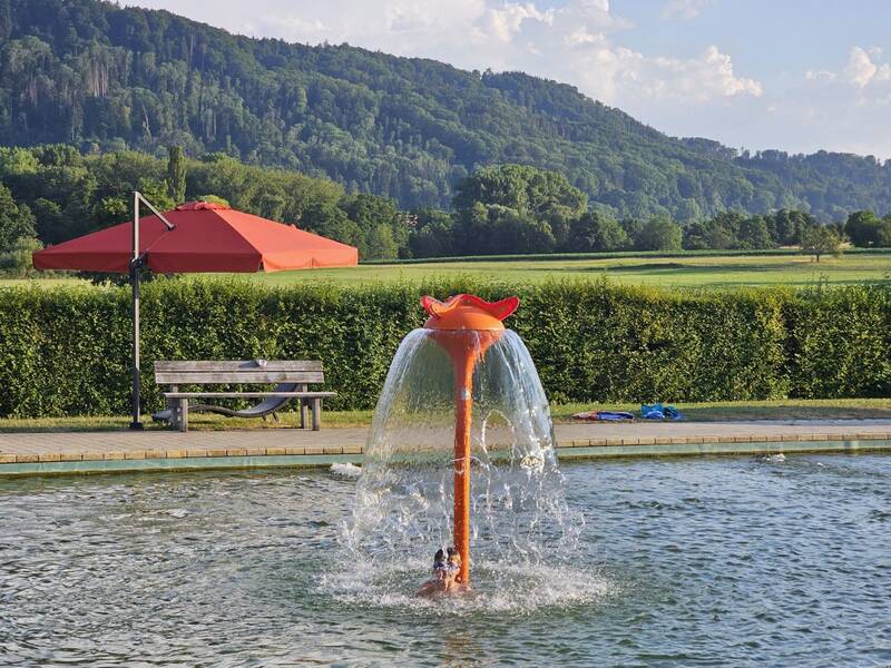 Ein oranges Wasserspiel in Form eines Pilzes sprüht Wasser in ein Becken vor einer grünen Hecke und einem roten Sonnenschirm, mit einem bewaldeten Hügel im Hintergrund.