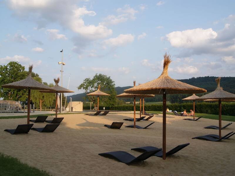 Strandbereich mit Sand, mehreren Sonnenliegen und strohgedeckten Sonnenschirmen, umgeben von grüner Landschaft unter leicht bewölktem Himmel.
