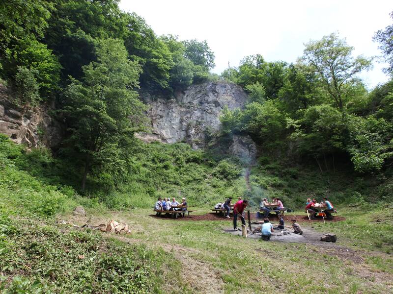 Eine Gruppe von Menschen sitzt an Tischen in einem von Bäumen umgebenen Naturgebiet, während in der Mitte ein Lagerfeuer brennt.