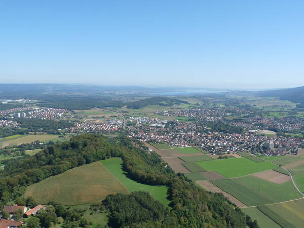 Eine weite Landschaft mit Wäldern, Feldern und einer Siedlung erstreckt sich bis zum Horizont, mit klarem Himmel darüber.