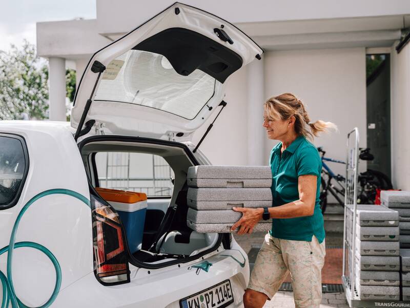 Eine Frau in einem türkisfarbenen Poloshirt lädt einen Stapel grauer Boxen in den Kofferraum eines weißen Autos.