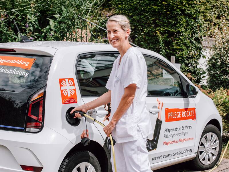 Eine Person in weißer Kleidung lächelt, während sie ein Elektroauto mit einer Ladestation verbindet.