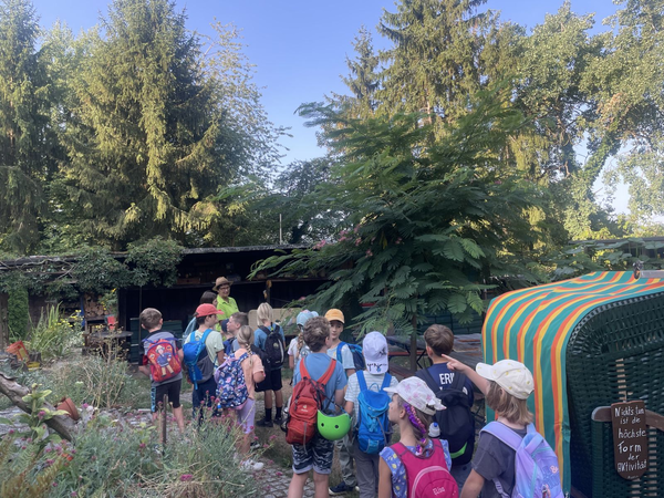 Stadtimkerin - Eine Gruppe von Kindern mit Rucksäcken steht im Freien vor einer Imkerin in grüner Kleidung unter Bäumen und neben einem bunten Strandkorb.