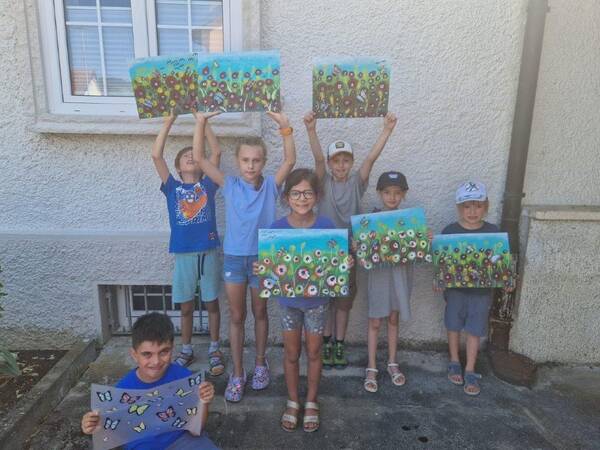 Eine Gruppe von sieben Kindern steht vor einer weißen Wand und hält farbenfrohe Gemälde mit Blumen- und Schmetterlingsmotiven hoch.