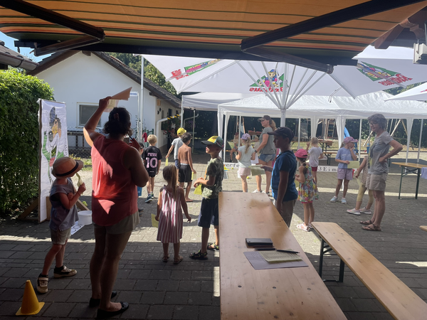 Auf dem Bild sind mehrere Erwachsene und Kinder in einem Außenbereich mit Pavillons und Bänken zu sehen, die an einem sonnigen Tag zusammenstehen, wobei einige Hüte tragen.