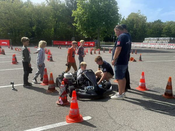 Eine Gruppe von Kindern und Erwachsenen steht auf einem Parkplatz um ein Kart herum, das von Pylonen umgeben ist.