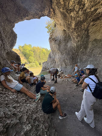 Eine Gruppe von Menschen, darunter viele Kinder, sitzt und steht in einer felsigen Höhle, während ein Erwachsener ihnen etwas zeigt.