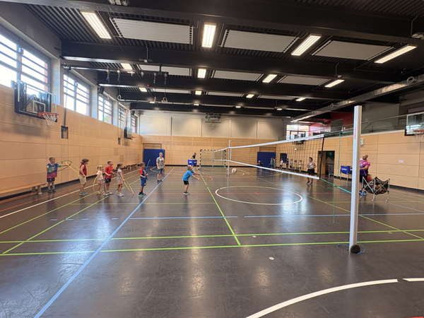 In einer Sporthalle spielen Kinder mit Reifen und Bällen, während Erwachsene am Rand stehen.