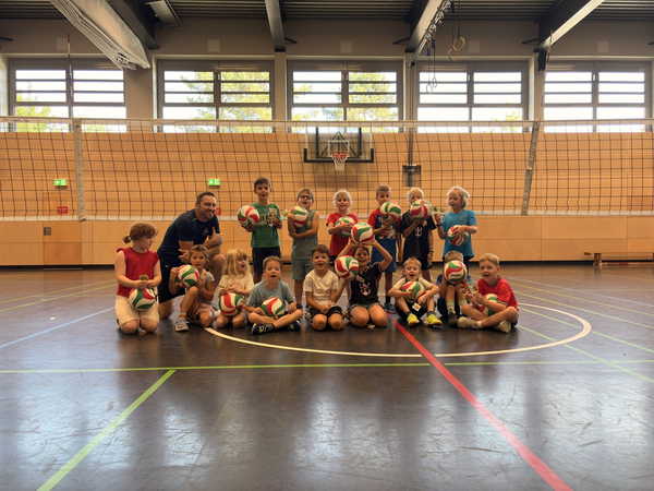 Eine Gruppe von Kindern und einem Erwachsenen posiert in einer Sporthalle vor einem Volleyballnetz mit Volleyballbällen.