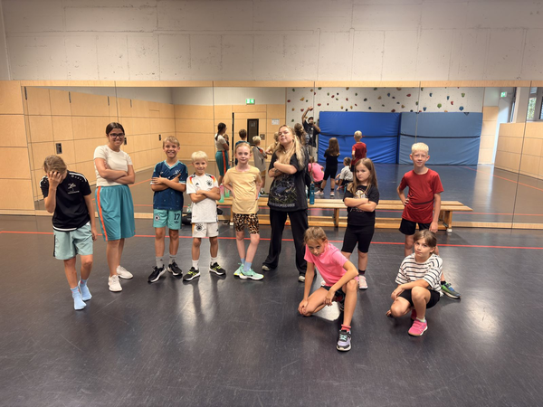 Eine Gruppe von Kindern und Jugendlichen steht und sitzt lässig in einem Raum mit spiegelverkleideten Wänden und Sportmatten im Hintergrund.