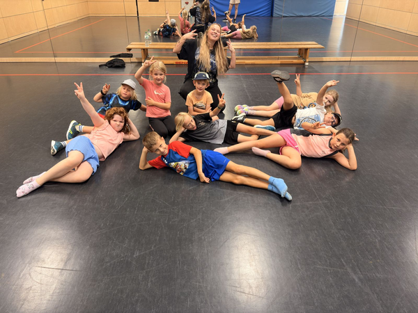 In einem Gymnastikraum posiert eine Gruppe fröhlicher Kinder mit einer lächelnden Erwachsenen zusammen auf dem Boden.