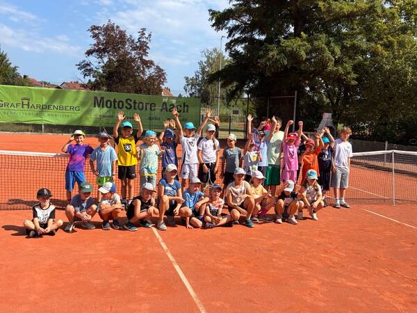 Eine Gruppe von Kindern steht fröhlich auf einem Tennisplatz, einige sitzen vor dem Netz und tragen bunte Sommerkleidung und Kappen.