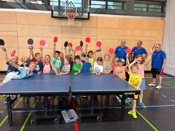 Eine Gruppe fröhlicher Kinder und Erwachsener steht um einen Tischtennistisch und hält farbige Tischtennisschläger in die Luft.