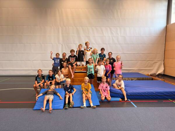 Eine Gruppe von Kindern und einem Erwachsenen sitzt und steht auf Turnmatten und einem Kasten in einer Sporthalle und lächelt in die Kamera.