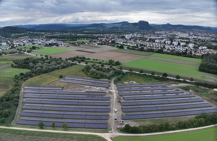 Das Bild zeigt ein großes Solarfeld vor einer Stadtlandschaft mit Feldern, Wohngebieten und Hügeln im Hintergrund unter einem bewölkten Himmel.