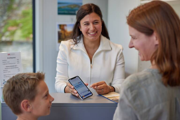 Eine Frau mit weißer Jacke zeigt einer Mutter und ihrem Sohn an einem Schalter ein Smartphone mit Informationen.
