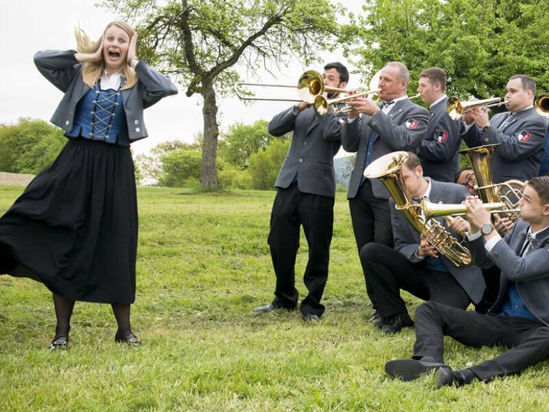 Eine Frau in traditioneller Tracht hält sich lachend die Ohren zu, während eine Gruppe von Musikern im Freien enthusiastisch Blasinstrumente spielt.
