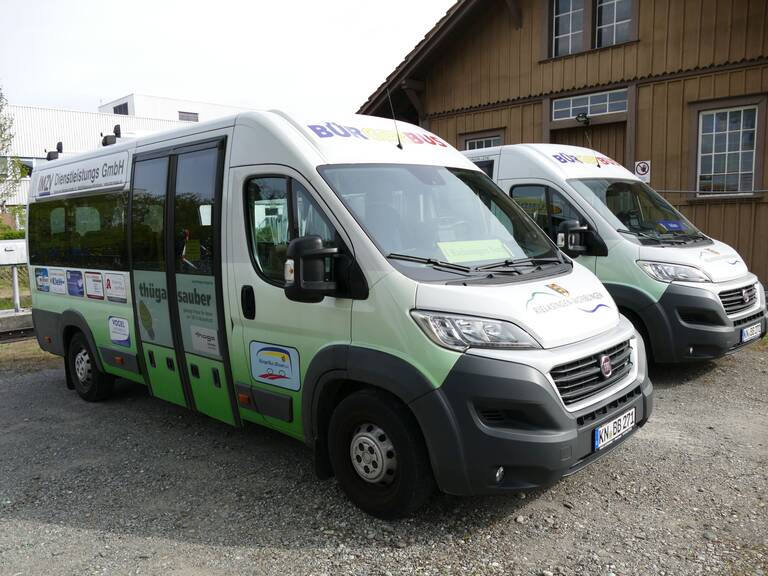 Zwei grün-weiße Kleinbusse mit der Aufschrift "Bürgerbus" stehen nebeneinander auf einem Platz vor einem Gebäude.