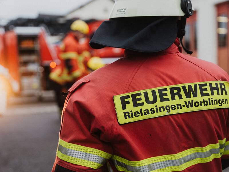 Ein Feuerwehrmann in roter Einsatzkleidung mit der Aufschrift "Feuerwehr Rielasingen-Worblingen" steht im Vordergrund, während andere Feuerwehrleute und ein Feuerwehrauto im Hintergrund unscharf zu sehen sind.