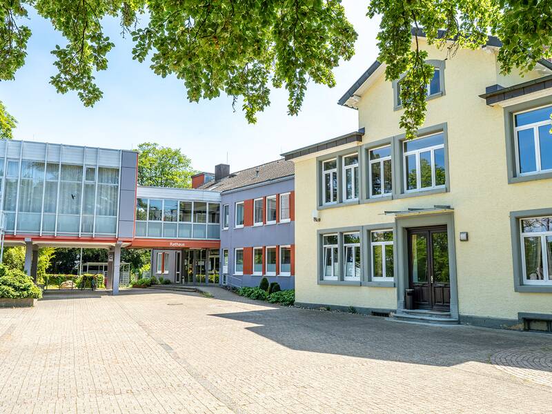 Ein modernes Gebäude mit gelber Fassade und großen Fenstern steht neben einem grauen Anbau, verbunden durch einen gläsernen Übergang, umgeben von grünen Bäumen.